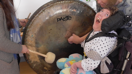 A young lady in a wheelchair is wowed by the sound of a large tam-tam brought to life by a soft beater